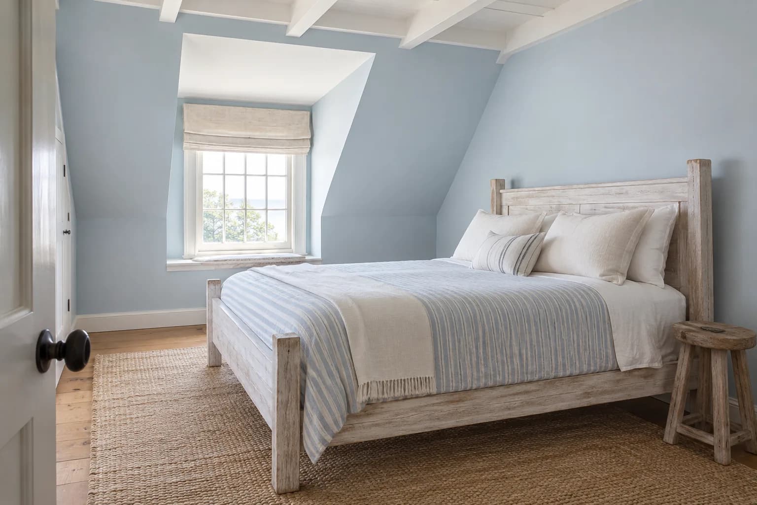 Soft blue attic bedroom with sloped walls, white ceiling beams, a whitewashed bed, and striped bedding