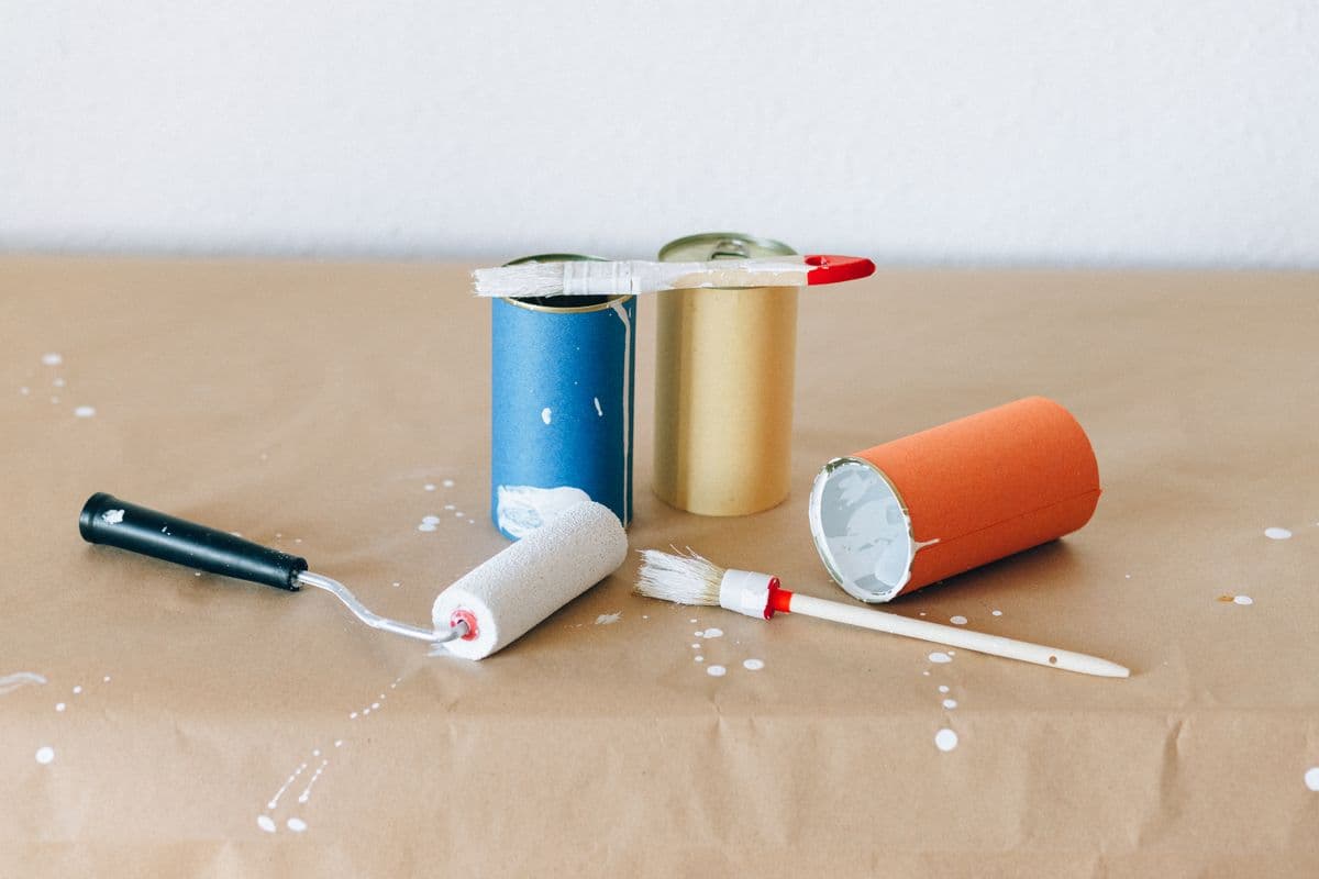 Paint cans, a roller, and a paint brush on a paper-covered table with white paint splatters