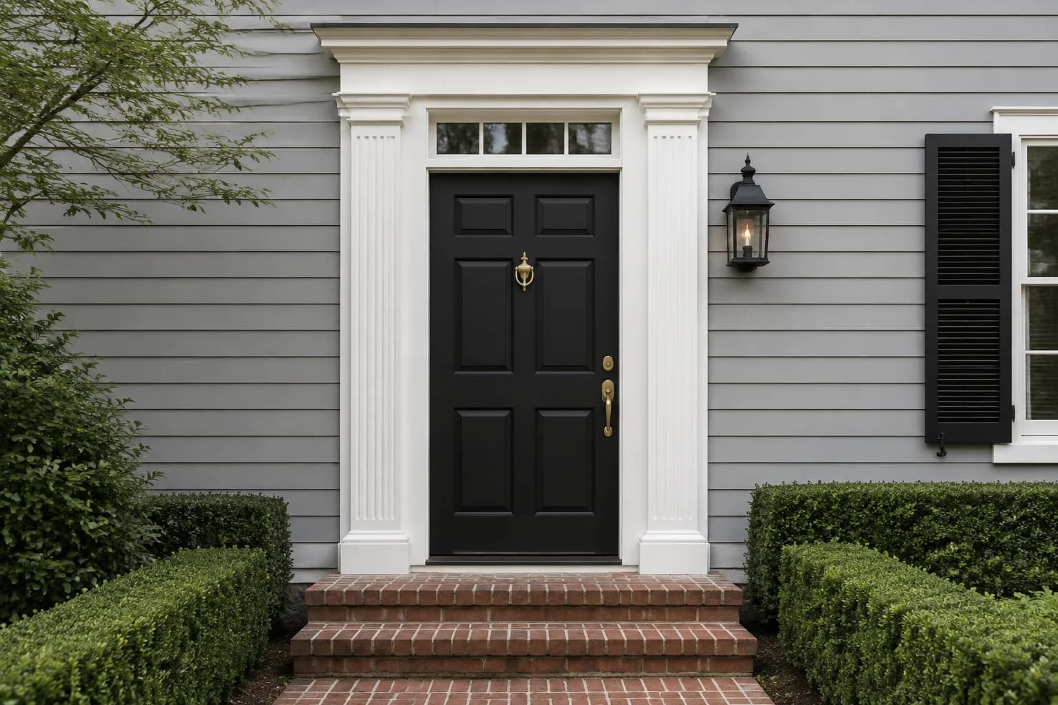 Black six-panel Colonial door with brass hardware, white pilasters, pale gray clapboard, and brick steps