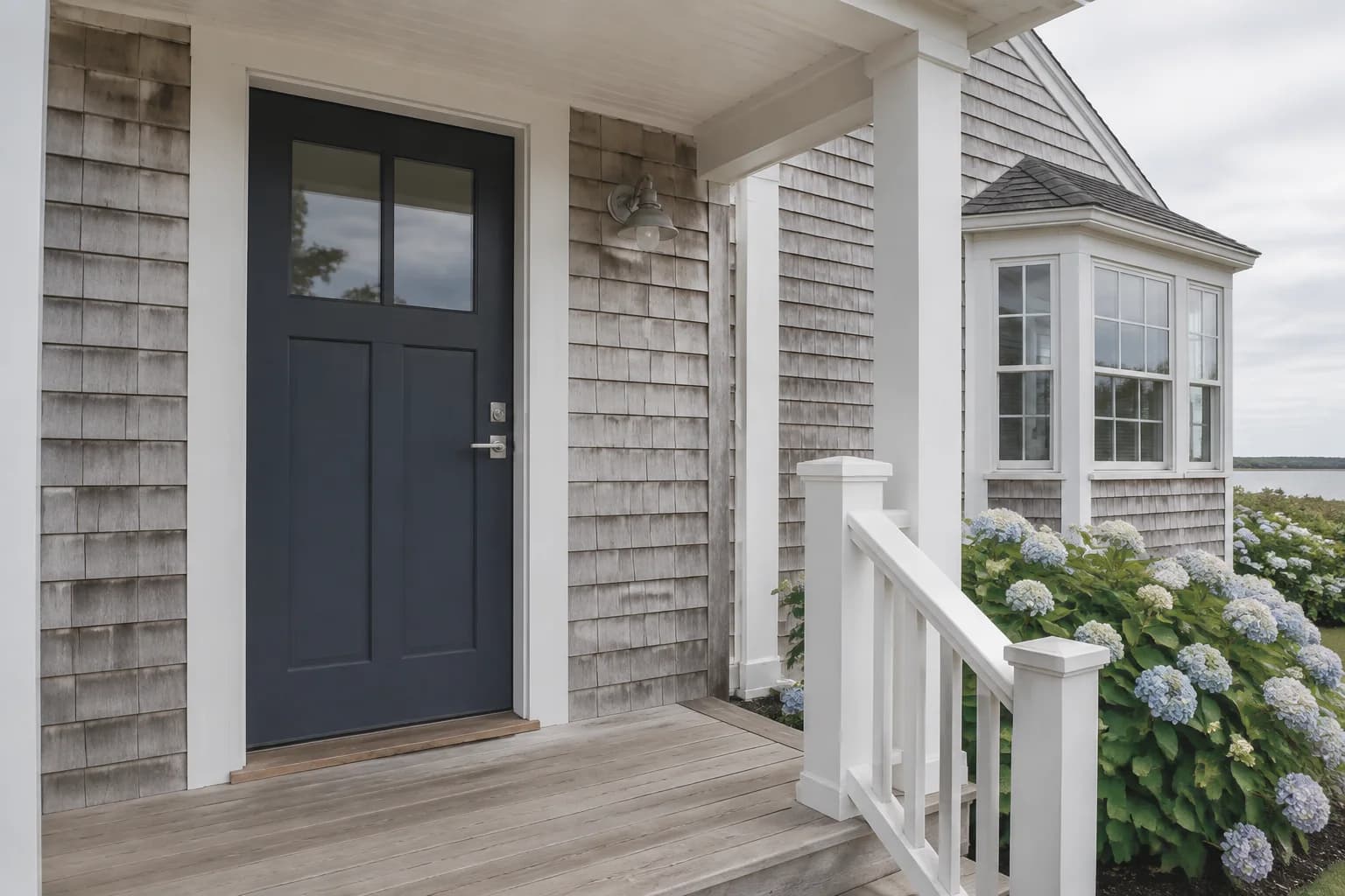 Navy glazed door on a gray-shingled coastal cottage with white porch railing and hydrangeas