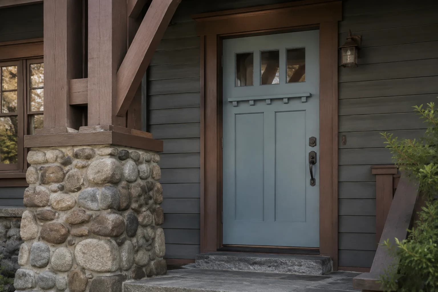 Slate blue Craftsman door with three upper windows and a dentil shelf beside dark gray siding and a river-stone porch pier