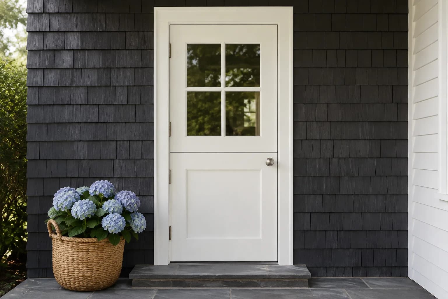 Closed soft white Dutch door with four upper glass panes, dark gray shingle siding, and a hydrangea basket
