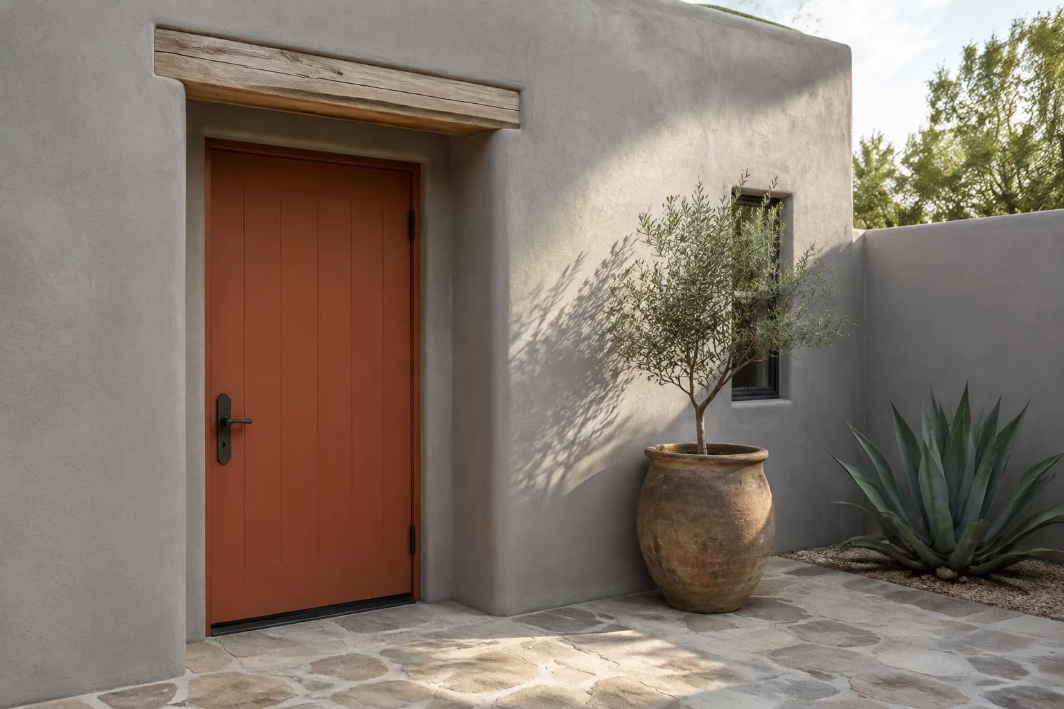 Terracotta plank door recessed into gray stucco beside a weathered clay planter, olive tree, and stone courtyard