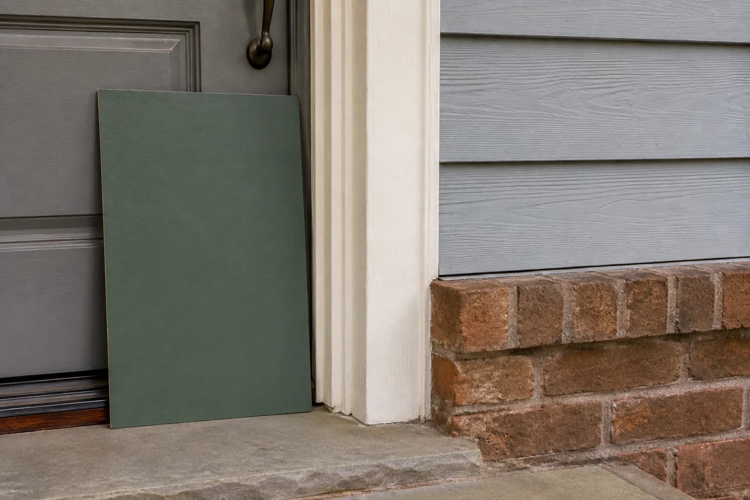 Large green paint sample board at a doorway beside off-white trim, blue-gray siding, warm brick, and a stone threshold