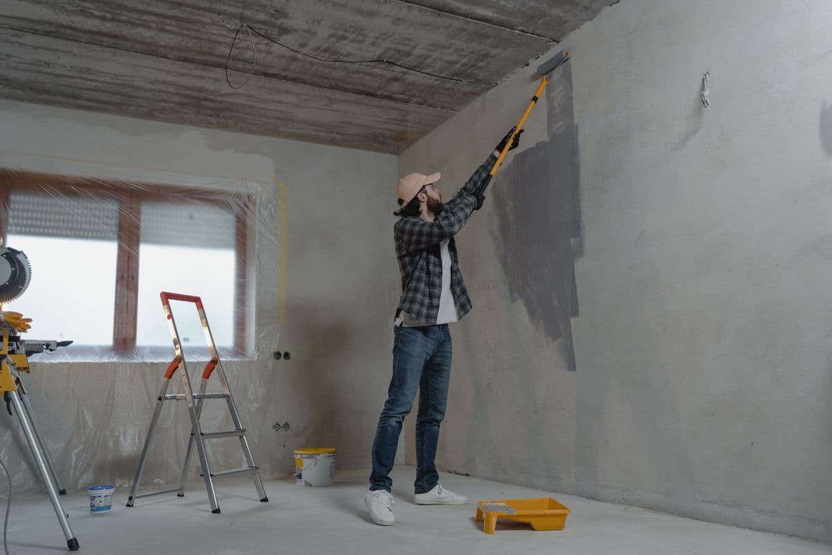 Man rolling gray paint onto a bare interior wall, with the fresh patch standing out from the unpainted surface